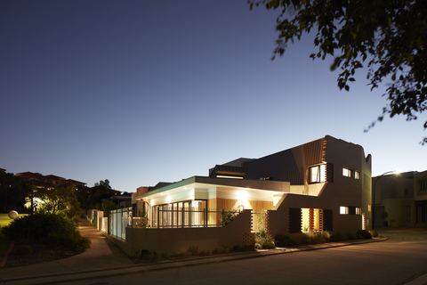 The City Beach house at dusk, its dark upper storey cantilevered over a lit ground floor and garden